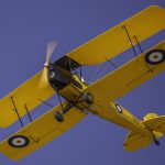 A yellow biplane with RAF roundels on the wings flying against a clear blue sky, captured in mid-air with its propeller in motion.